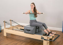Load image into Gallery viewer, A woman exercising on a contour sitting box, using a maple dowel and attached Pilates foot straps.
