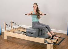 Load image into Gallery viewer, A woman exercising on a contour sitting box, using a maple dowel and attached Pilates foot straps.
