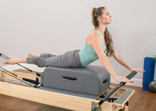 Load image into Gallery viewer, A woman leveraging a contour sitting box for enhanced stability while on a reformer.
