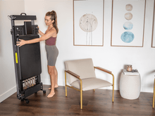 Load image into Gallery viewer, A woman is storing a Metro-IQ Pilates Reformer vertically on a wall.

