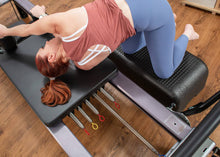 Load image into Gallery viewer, A woman stretching out on a Pilates reformer, with signature springs prominently displayed.
