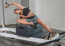 Load image into Gallery viewer, Woman stretching on a sitting box, using Pilates foot straps attached to a reformer.

