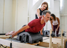 Load image into Gallery viewer, A man engaging in a workout on a Pilates reformer under the guidance of an instructor.
