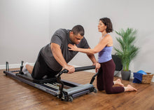 Load image into Gallery viewer, Female instructor guiding a man on how to use an Allegro Stretch Pilates Reformer.
